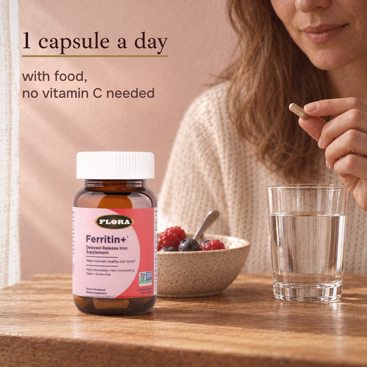 A woman holds one Ferritin+ capsule beside the bottle, a bowl of berries and a glass of water: 1 capsule a day, with food, no vitamin C needed